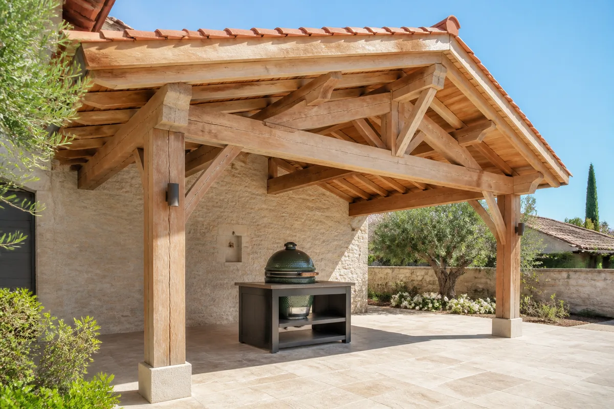 Auvent en charpente bois couvert de tuiles abritant un barbecue céramique sur une terrasse en pierre