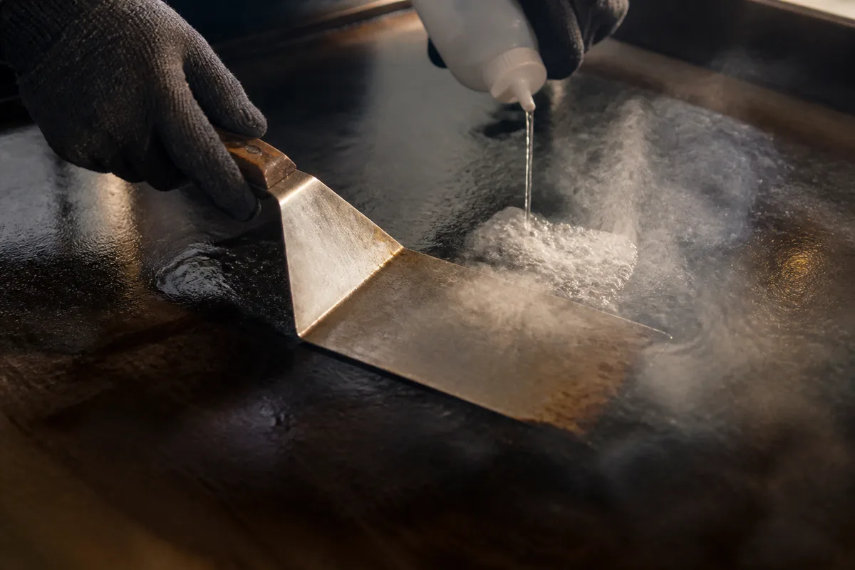 Mains gantées passant une spatule métallique sur une plaque de cuisson en acier culottée brun sombre, vapeur légère au contact de l’eau