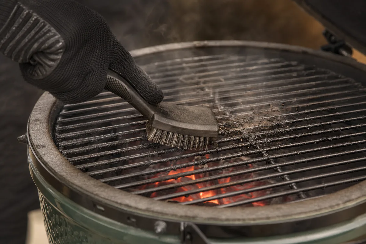 Brossage d&rsquo;une grille de cuisson chaude au-dessus du foyer d&rsquo;un kamado, résidus qui se détachent sous la brosse