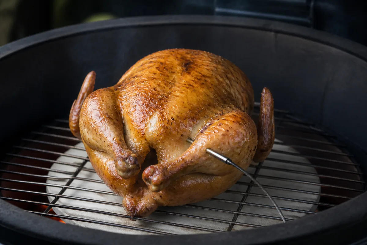 Poulet entier doré posé sur la grille d’un barbecue en céramique ouvert, braises rougeoyantes visibles sous le déflecteur